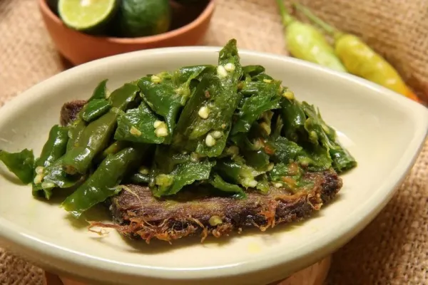 A popular Padangnese dish of dried beef jerky topped with green chili. Plated on a oval ceramic plate. In the background are a bowl of limes and chili peppers.