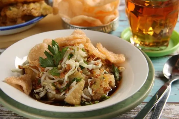 Kupat Tahu Magelang, a signature dish from Magelang, Central Java. It consists of rice cake (kupat or ketupat), bean curd (tahu), vegetable fritter, cabbage and beansprouts in sweet soy broth enriched with ground peanuts. Served on a white ceramic plate underlined with a green plate. A pair of spoon and fork is placed next to it. A tea in a glass is placed on the upper right hand side. An extra serving of crispy crackers and vegetable fritters are placed nearby. The meal is set on a rustic wooden table painted with bright colours of white and blue.