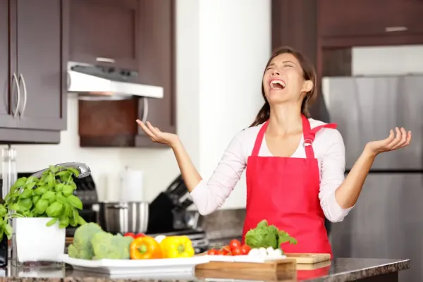 A frustrated woman in a red apron throws her hands up in the kitchen, surrounded by vegetables.