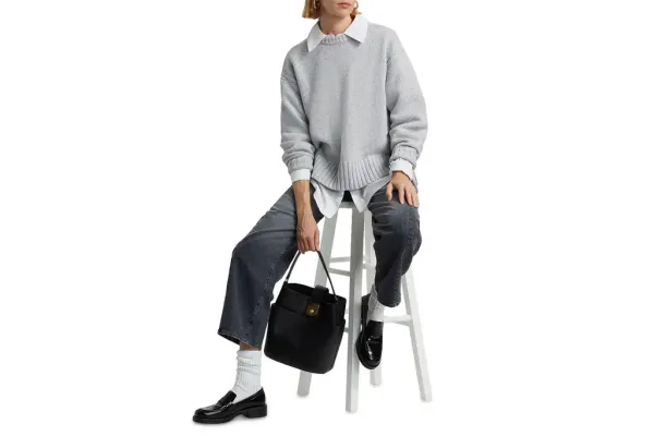 A person wearing a grey cashmere sweater over a white collared shirt, washed black jeans, white socks, and black loafers, sitting on a white stool and holding a black leather bucket bag.