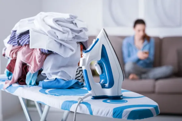 A woman lounges on a couch, texting on her phone, while a pile of laundry and an iron sit on an ironing board in the foreground.