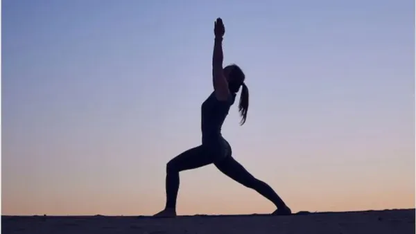 A woman performing Virabhadrasana (Warrior Pose) outdoors against a clear sky, with arms raised and body in a strong, balanced stance.