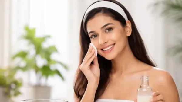 A smiling woman with a white headband gently cleansing her face with a cotton pad while holding a skincare bottle in a bright, natural indoor setting.