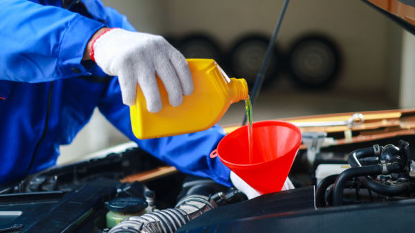 A mechanic in blue coveralls and white gloves pouring oil into a car engine with a red funnel