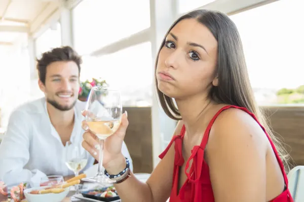 A woman with a bored expression holds a glass of white wine while a man smiles in the background, implying a bad date.