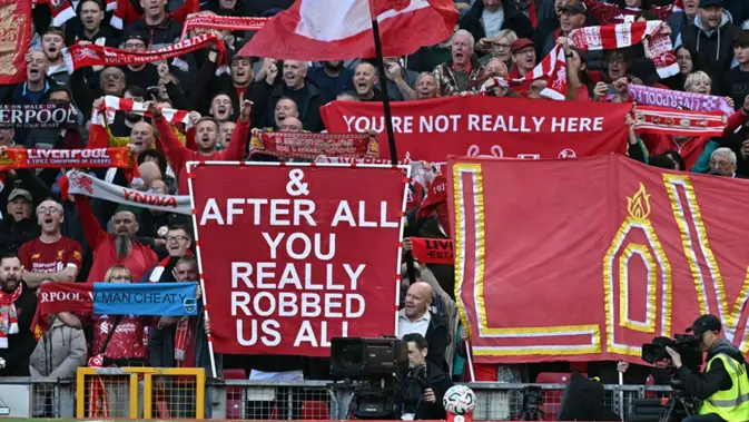 Penggemar Liverpool memegang poster yang mengejek skandal keuangan Manchester City saat kru televisi bekerja di tepi lapangan menjelang pertandingan Liga Inggris antara Liverpool dan Manchester City di Anfield di Liverpool, barat laut Inggris pada 11 Oktober 2026. Manchester City menghadapi Anfield yang bermusuhan pada hari Minggu ketika pemimpin Premier League itu menghadapi rival sengitnya Liverpool dalam pertandingan pertama mereka sejak dinyatakan bersalah karena melanggar lebih dari 100 aturan keuangan. (Paul ELLIS/AFP)