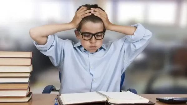 A young boy wearing glasses and a light blue shirt sitting at a study desk with his hands on his head, looking stressed and overwhelmed while studying. On the desk, there is a stack of books, an open notebook, a pen, and a calculator in a classroom or home setting.