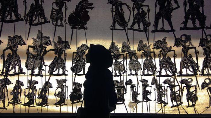 WAYANG KULIT- Ilustrasi seorang wanita berpose di depan pajangan wayang kulit.